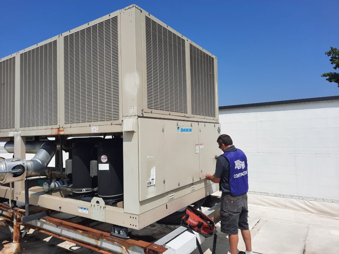 An SBS-NC technician at the control panel of a Daikin air-cooled chiller on a commercial rooftop, with the compressors and refrigerant piping visible